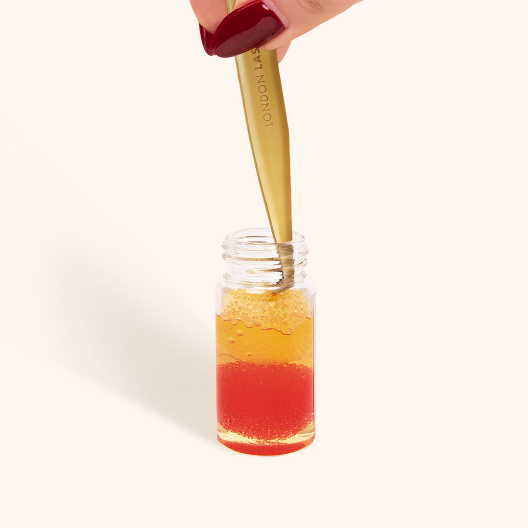 a close up of a bottle of glue remover for tweezers with a pair of gold lash tweezers being dipped into the liquid