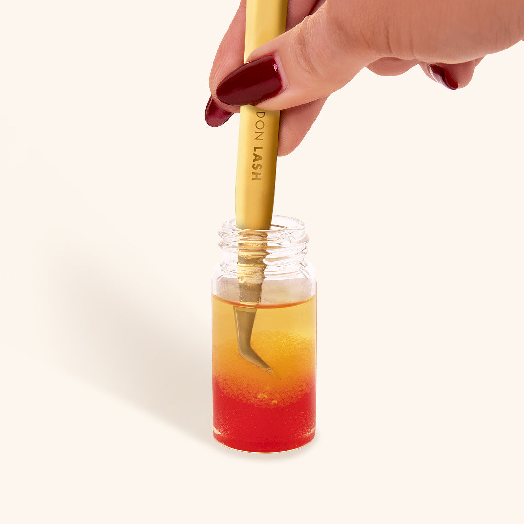 a close up of a bottle of glue remover for tweezers with a pair of gold lash tweezers being dipped into the liquid